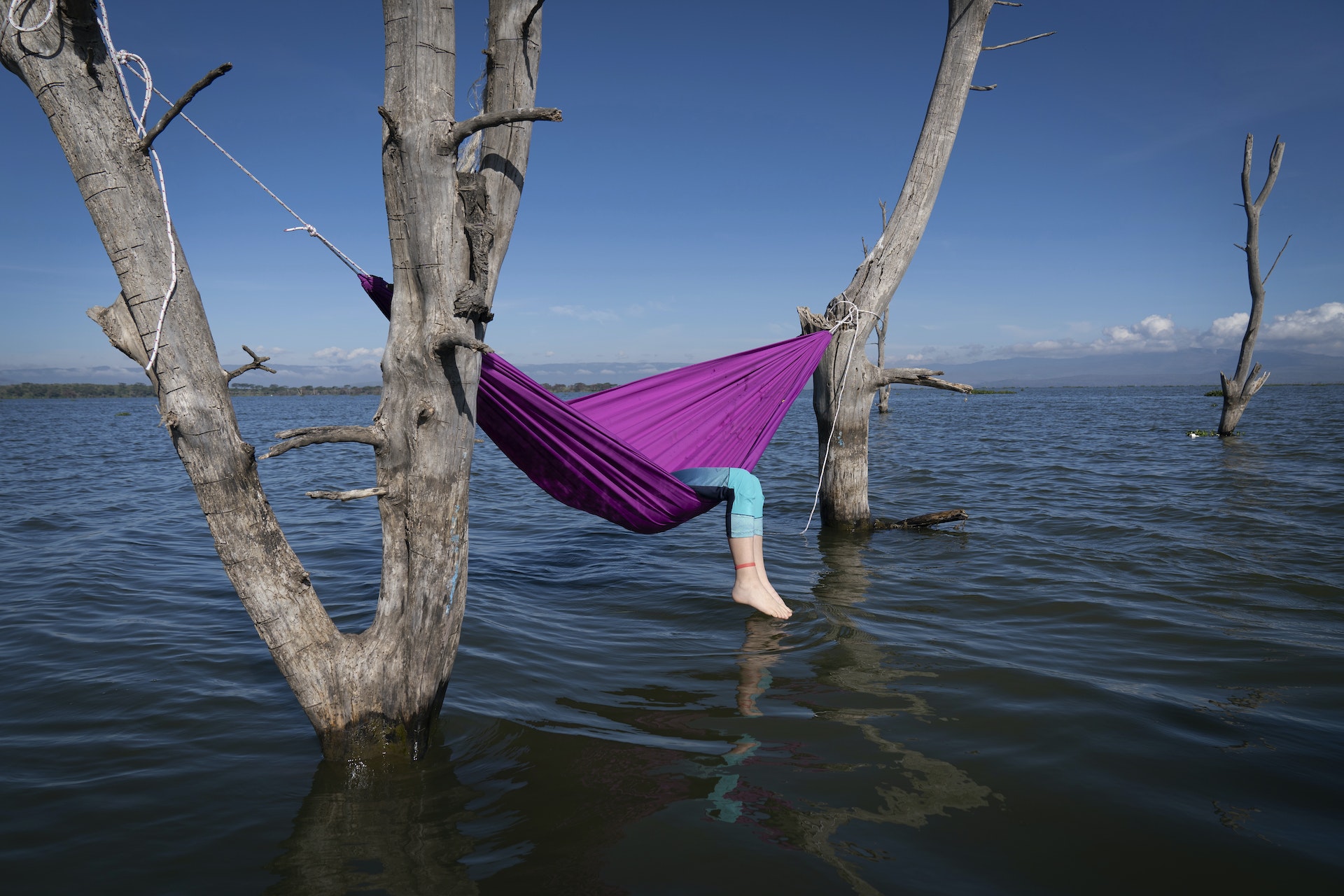 A person reclines in a hammock that is strung up between two pieces of wood over a lake